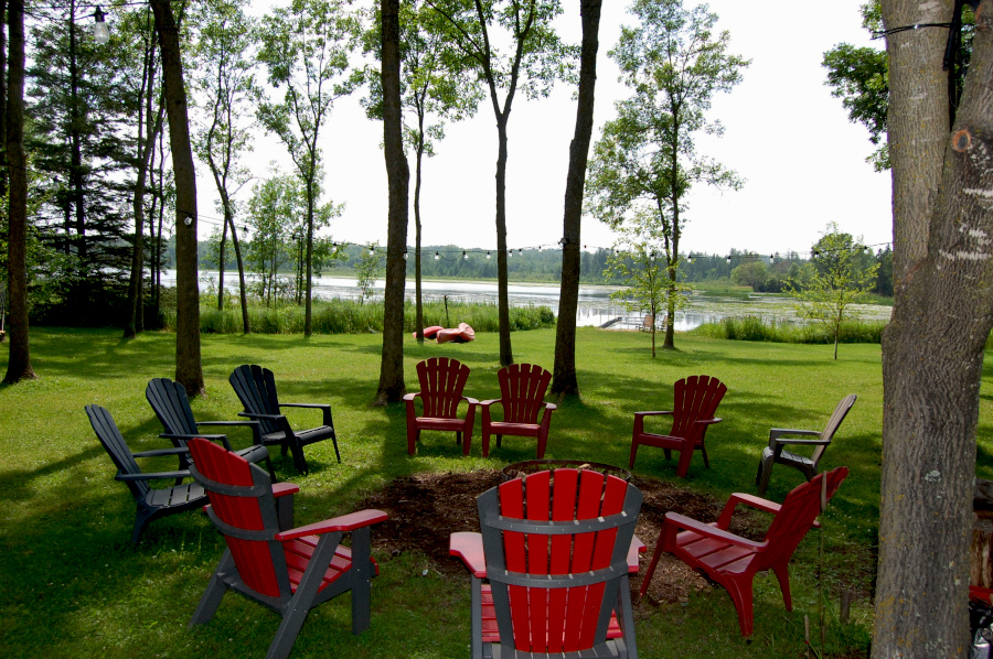 A large, grassy lakefront with a picturesque view of Bone Lake, a campfire surrounded by camp chairs.