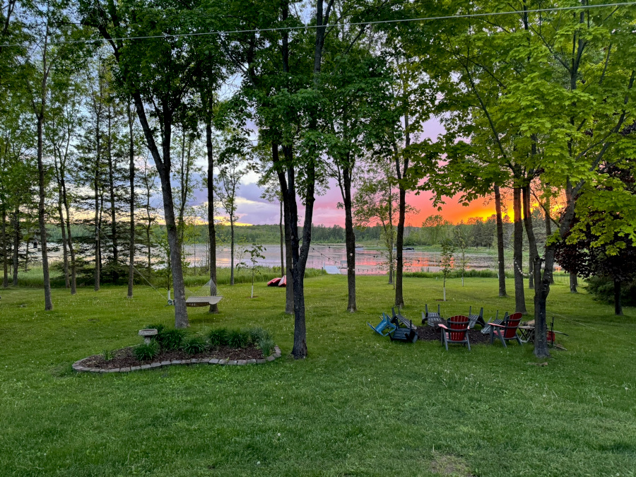 A grassy backyard overlooking a lake, with mature trees and a high canopy that provide an unobstructed West/Northwest view of the water and sunset. Adirondack chairs sit near a firepit, and the sky glows orange and pink at dusk.