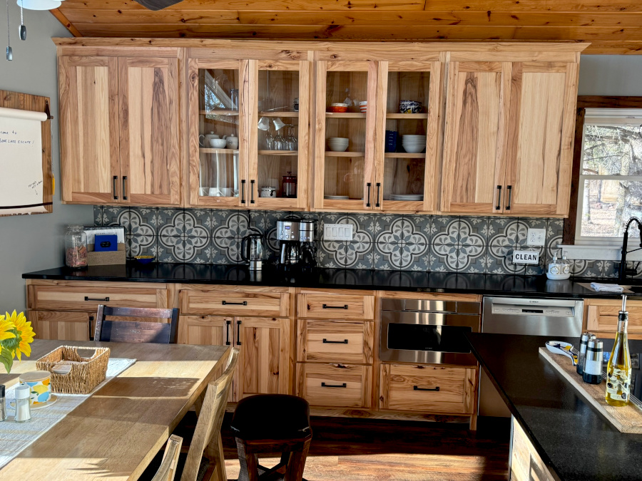 Hickory and carriage pine cabinetry hold dish ware and other kitchen essentials. Warm sunshine streams in on vast granite prep surfaces, a drip coffee maker, a large under mount sink.