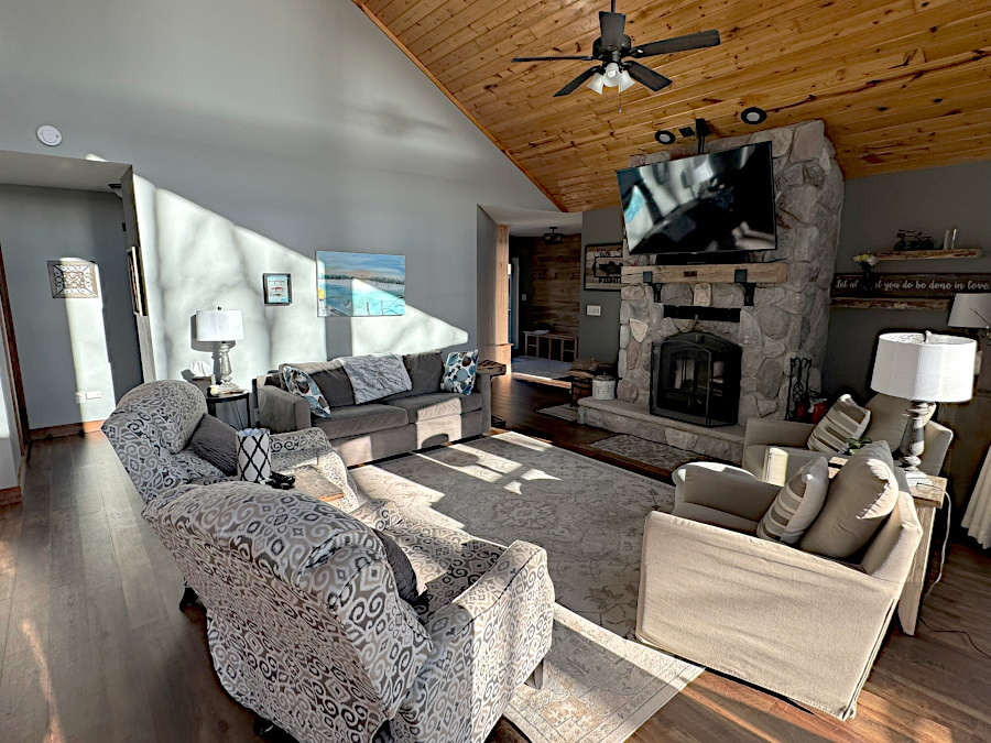Bright living room with vaulted wood ceilings, a large stone fireplace with a mounted TV, comfortable seating including sofas and armchairs, and sunlight streaming across the hardwood floor.