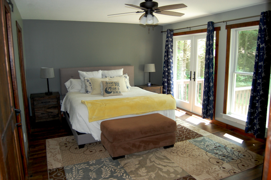 Primary bedroom with a king bed, patterned rug, matching nightstands, ceiling fan, and large windows with double doors leading to a private walkout deck.